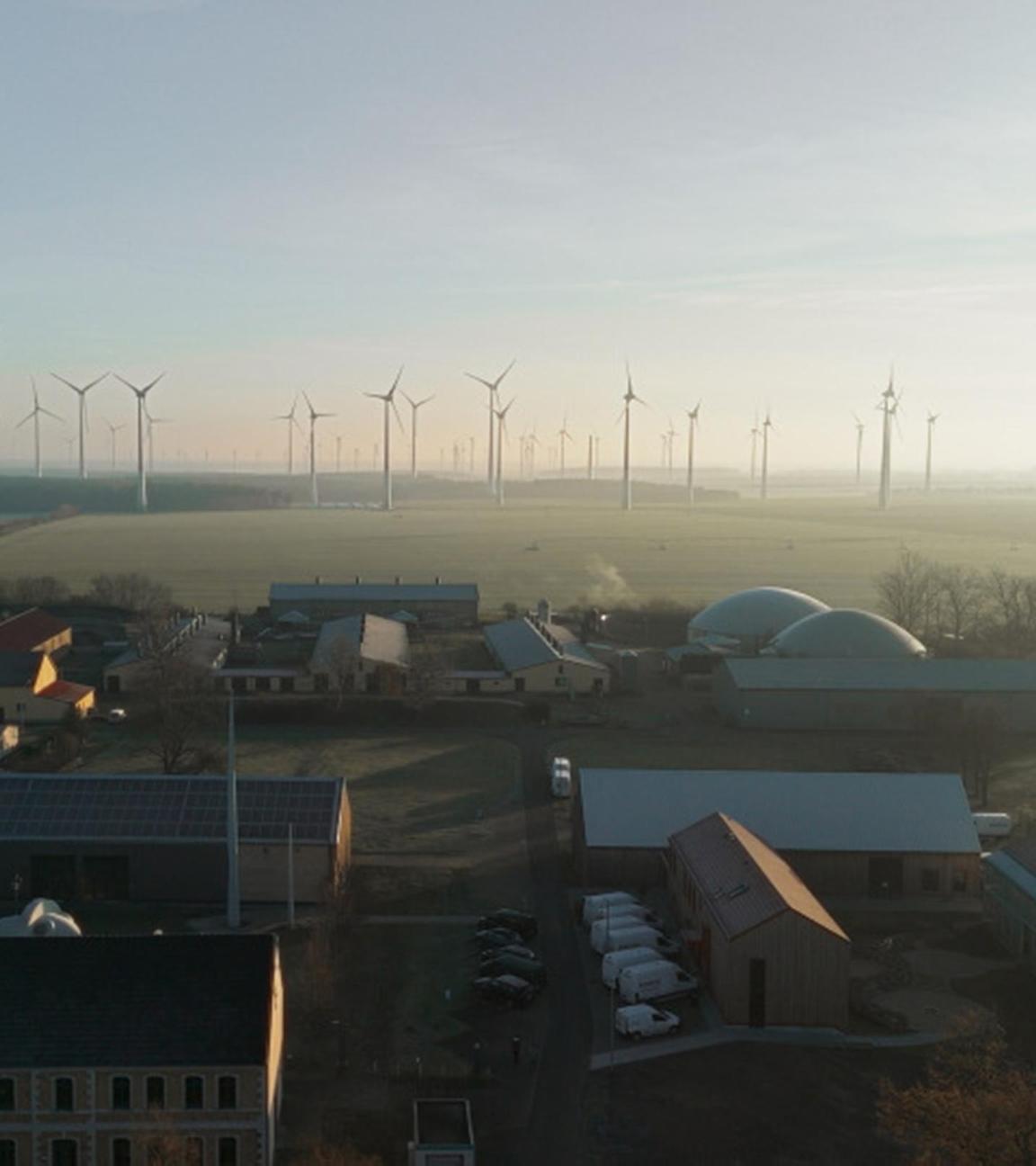 Luftaufnahme eines Dorfes, im Hintergrund sind Felder und Windräder zu sehen.