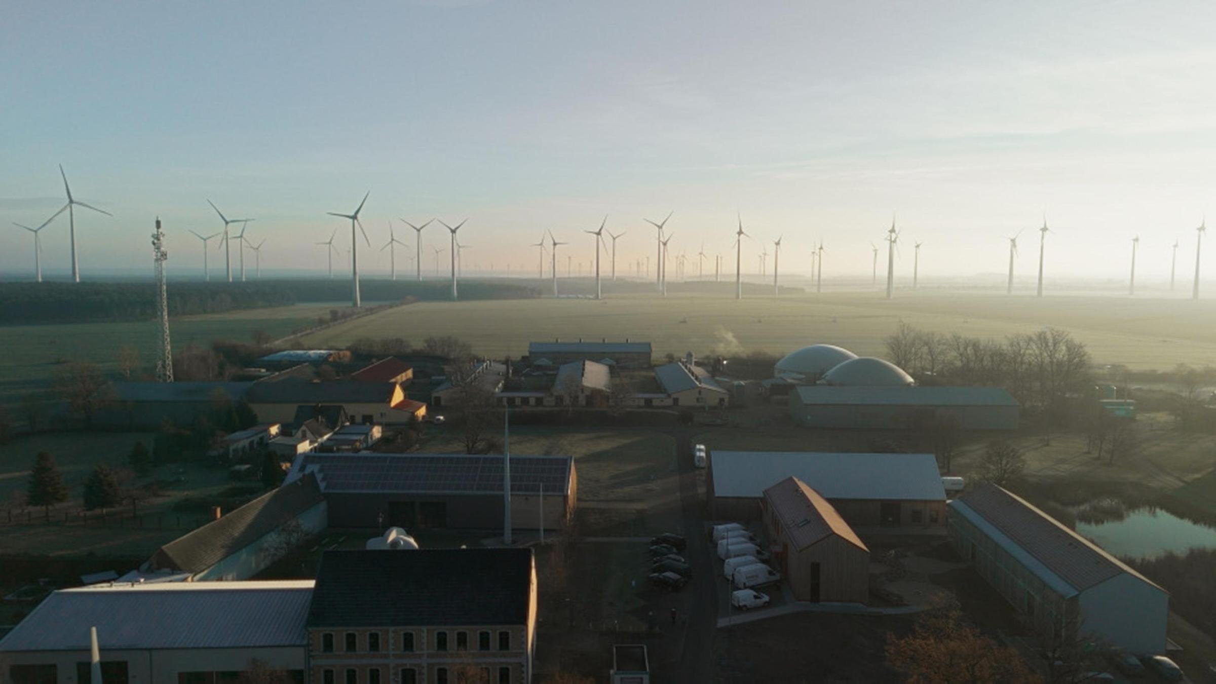 Luftaufnahme eines Dorfes, im Hintergrund sind Felder und Windräder zu sehen.