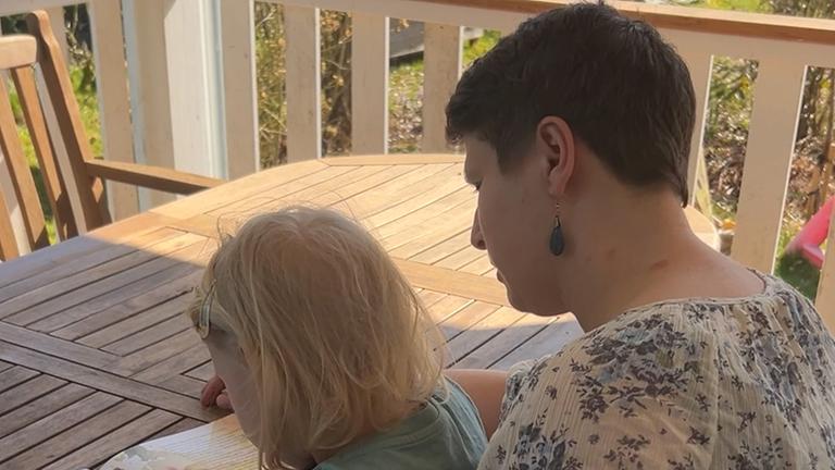 Eine Frau sitzt mit ihrer Tochter auf dem Schoß auf einer Terasse an einem Holztisch