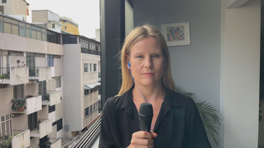 finnische Reporterin Maija Salmi berichtet aus Caracas, Venezuela. Sie trägt ein schwarzes Hemd und hält ein Mikrofon. Sie ist in einem Gebäude an einem Fenster und man kann die gegenüberliegenden Gebäude sehen.