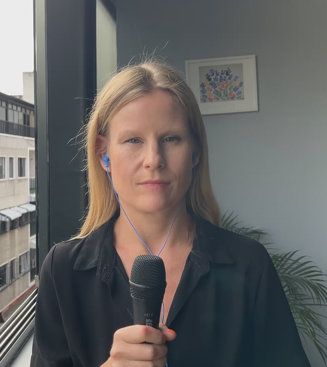 finnische Reporterin Maija Salmi berichtet aus Caracas, Venezuela. Sie trägt ein schwarzes Hemd und hält ein Mikrofon. Sie ist in einem Gebäude an einem Fenster und man kann die gegenüberliegenden Gebäude sehen.