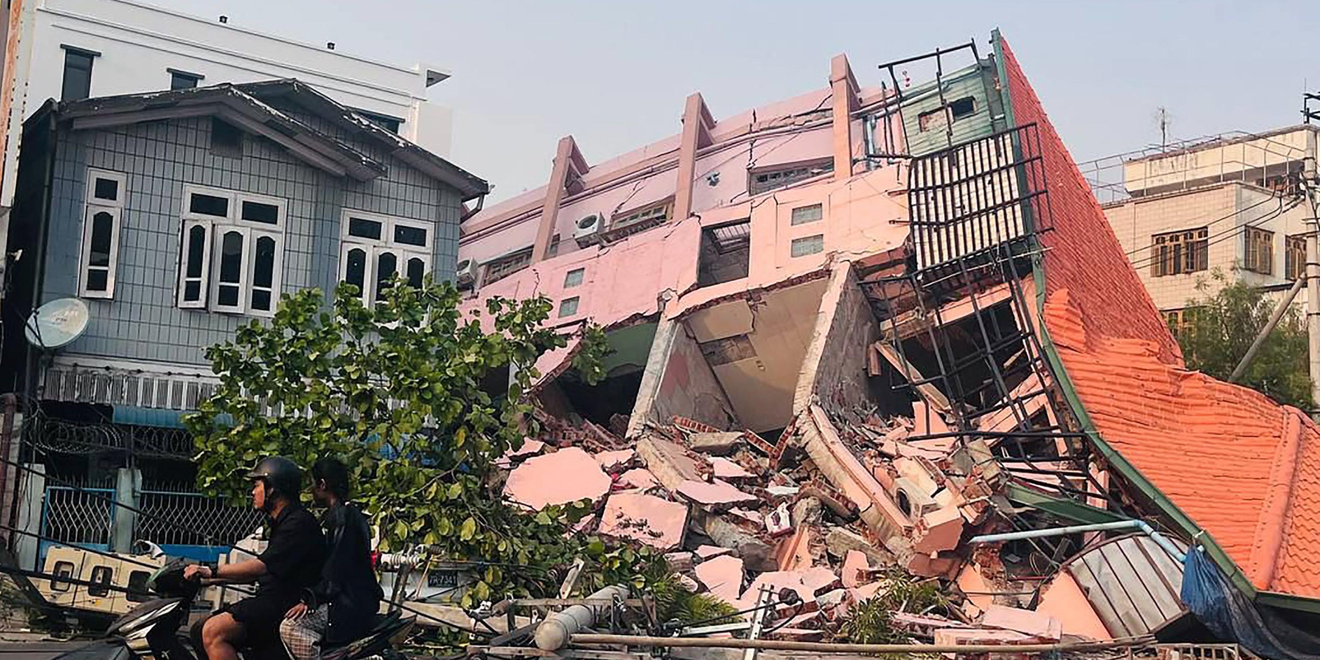 Menschen fahren am 28. März 2025 nach einem Erdbeben in Zentralmyanmar auf einem Motorrad an einem eingestürzten Gebäude in Mandalay vorbei.