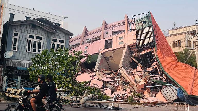Menschen fahren am 28. März 2025 nach einem Erdbeben in Zentralmyanmar auf einem Motorrad an einem eingestürzten Gebäude in Mandalay vorbei.