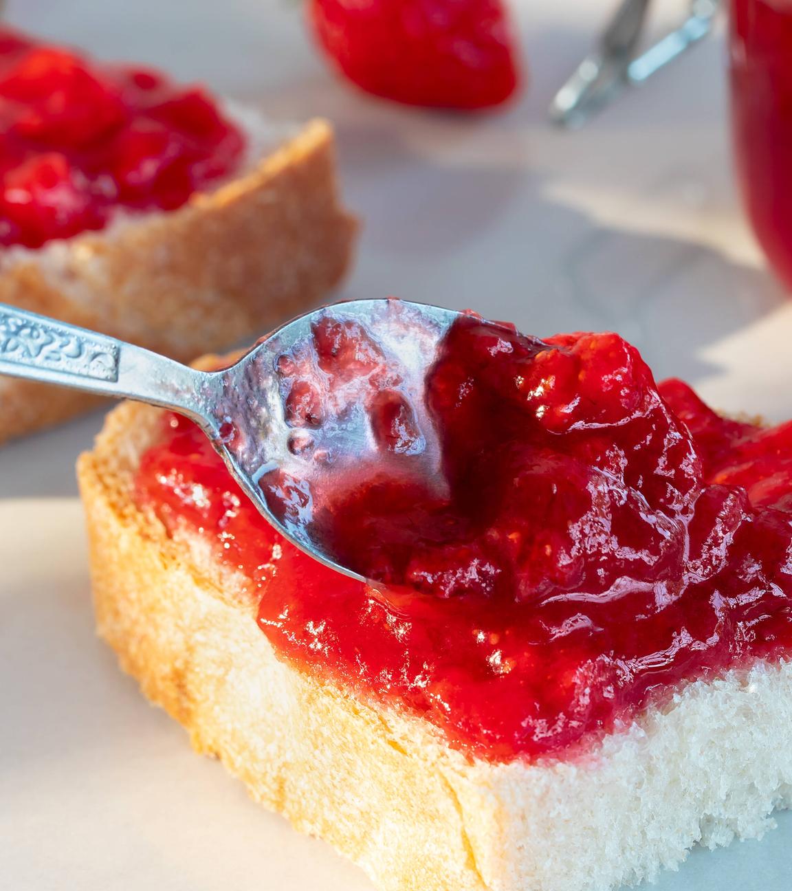 Auf einer halben Scheibe Brot wird mit einem Löffel Erdbeer-Fruchtaufstrich verteilt.