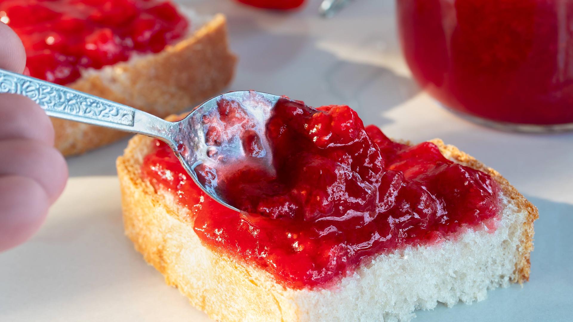 Auf einer halben Scheibe Brot wird mit einem Löffel Erdbeer-Fruchtaufstrich verteilt.