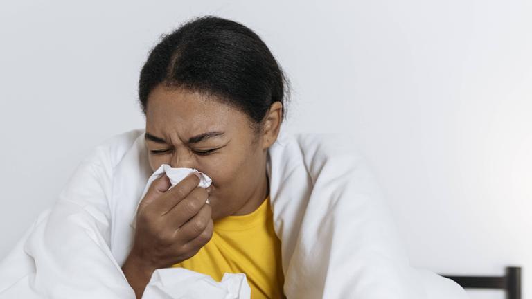 Symbolbild: Eine junge Frau sitzt auf dem Bett in eine Decke gehüllt und schnäuzt sich die Nase