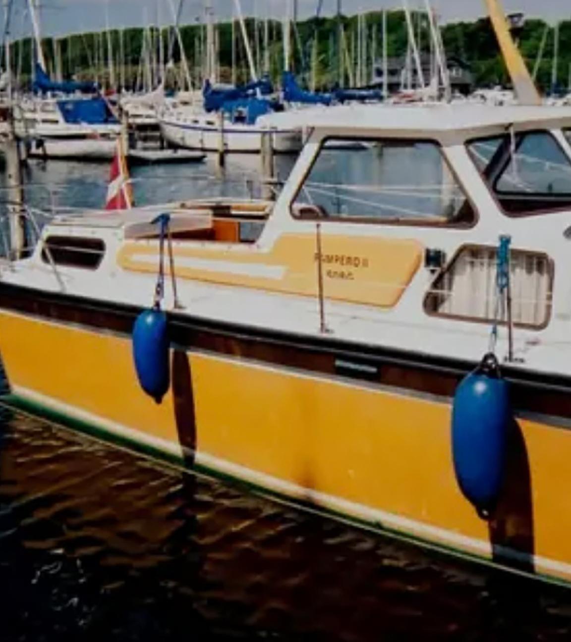 Ein weiß-gelbes Motorboot mit Kajüte liegt im Hafen. Das Boot trägt die dänische Flagge, im Hintergrund liegen weitere Segelboote.