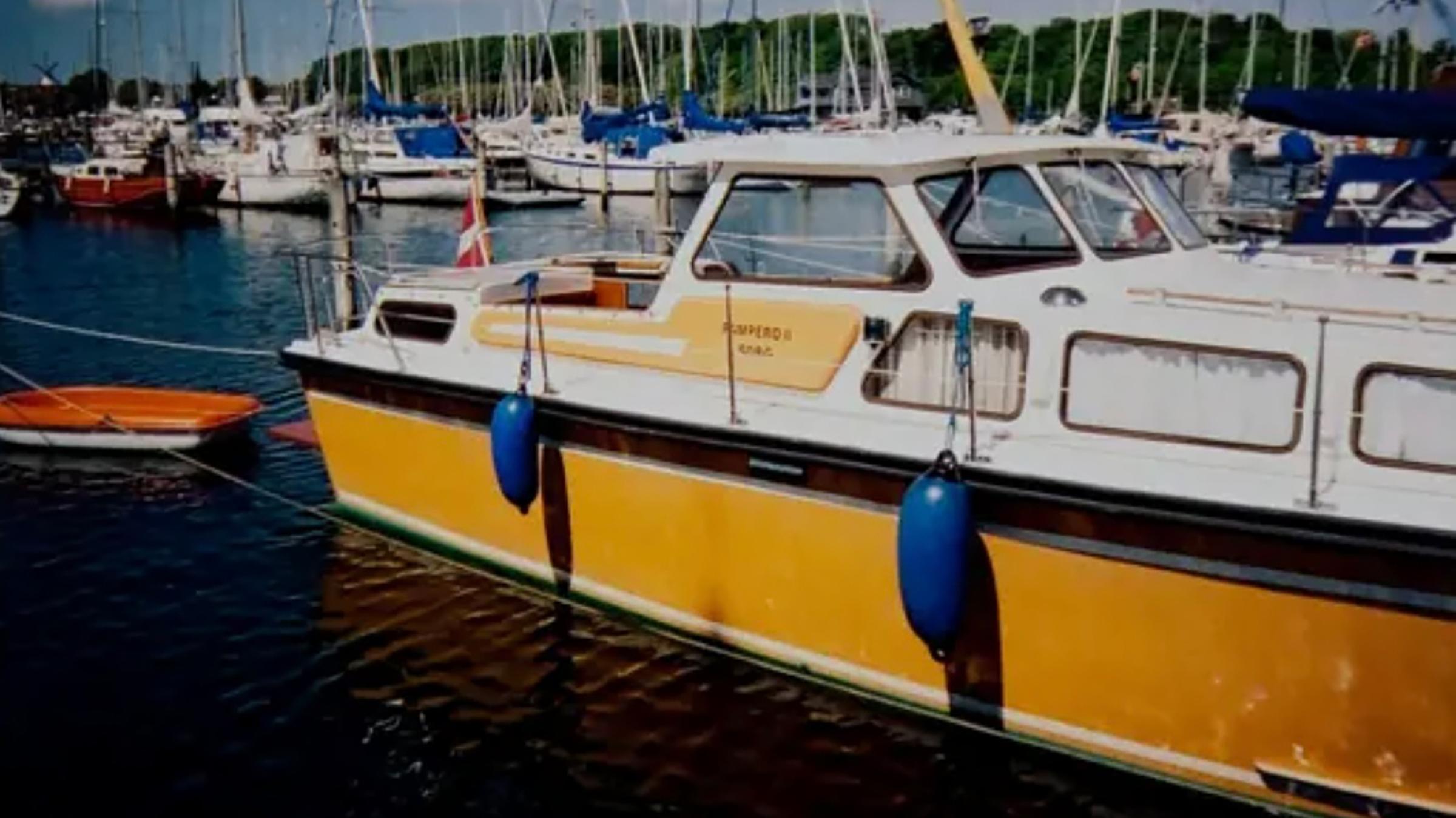 Ein weiß-gelbes Motorboot mit Kajüte liegt im Hafen. Das Boot trägt die dänische Flagge, im Hintergrund liegen weitere Segelboote.