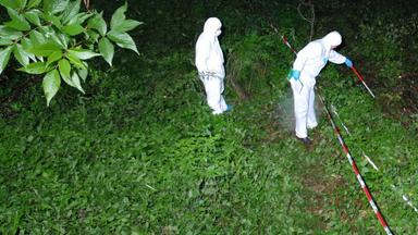 Zwei Ermittler in weißen Kriminaltechnikeranzügen durchsuchen nachts einen Haufen Äste in einem Wald. Der eine Ermittler tastet sich mit einem weiß-roten-Stock hinter einer Flatterbandabsperrung vor. Der andere Ermittler beobachtet von hinten die Szenerie. Die Markierung "Polizeifoto" liegt darüber.