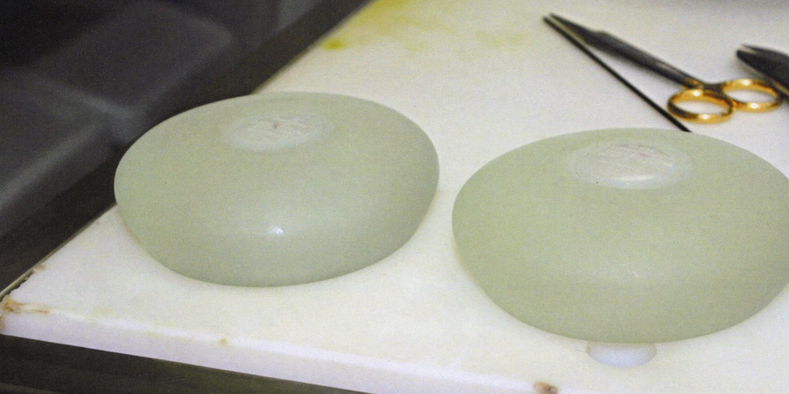 Zwei weiße, ovale Silikonkissen liegen auf einem weißen Marmortisch. Oberhalb des rechten Implantats liegt eine Operationsklammer.
