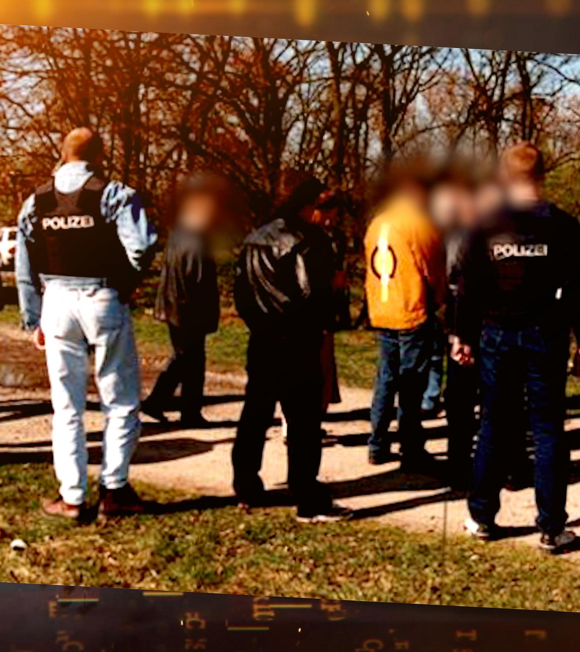 Mehrere Personen stehen auf einem schmalen Feldweg zwischen Wiese und Bäumen. Einige tragen Westen mit der Aufschrift "Polizei". Alle Personen stehen mit dem Rücken zur Kamera. Die Gesichter sind unkenntlich gemacht