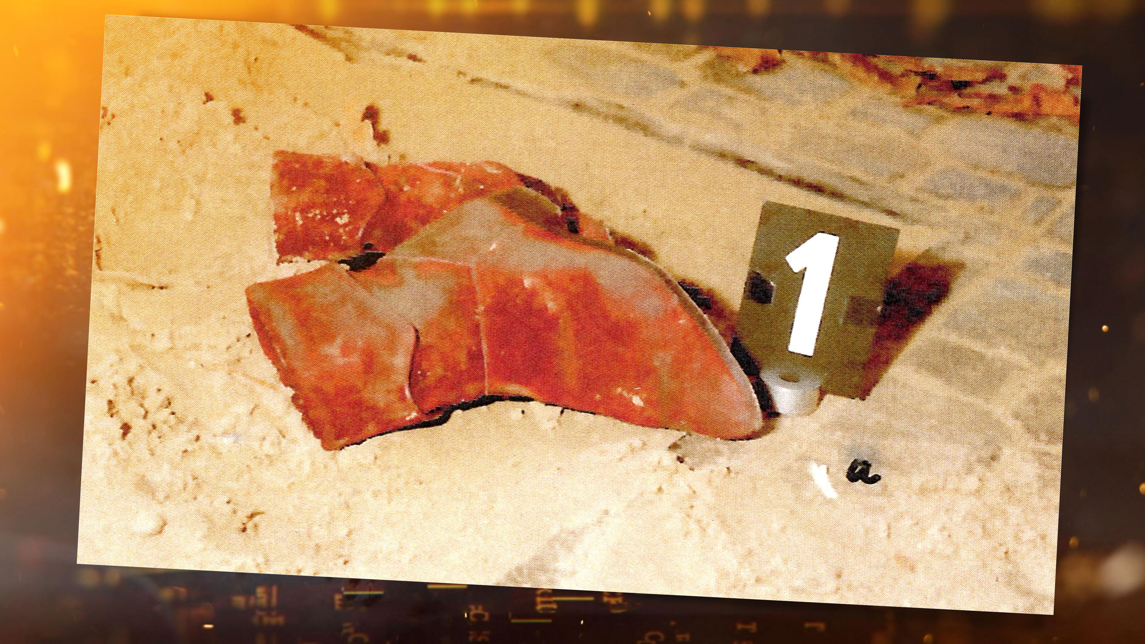 Tatortfoto: In einem Sandkasten liegt ein Paar rote Stiefel, daneben steht ein schwarzes Schild mit einer weißen Nummer "1". Den Sandkasten umgibt graues Kopfsteinpflaster, es ist teilweise ebenfalls mit Sand bedeckt. Oberhalb der Stiefel auf dem Kopfsteinpflasterst liegt, schemenhaft zu erkennen, ein Schal mit einer schwarze Tafel mit der weißen Nummer "4". An der Seite steht eine schwarze Tafel mit der Nummer "5" auf den Pflastersteinen.
