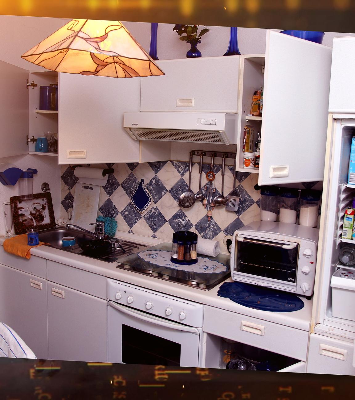 In einer weißen Einbauküche, die sehr aufgeräumt wirkt, sind Ober- und Unterschränke teilweise geöffnet. Im geöffneten Kühlschrank an der Seite sieht man verschiedene Getränke und Joghurtpackungen.