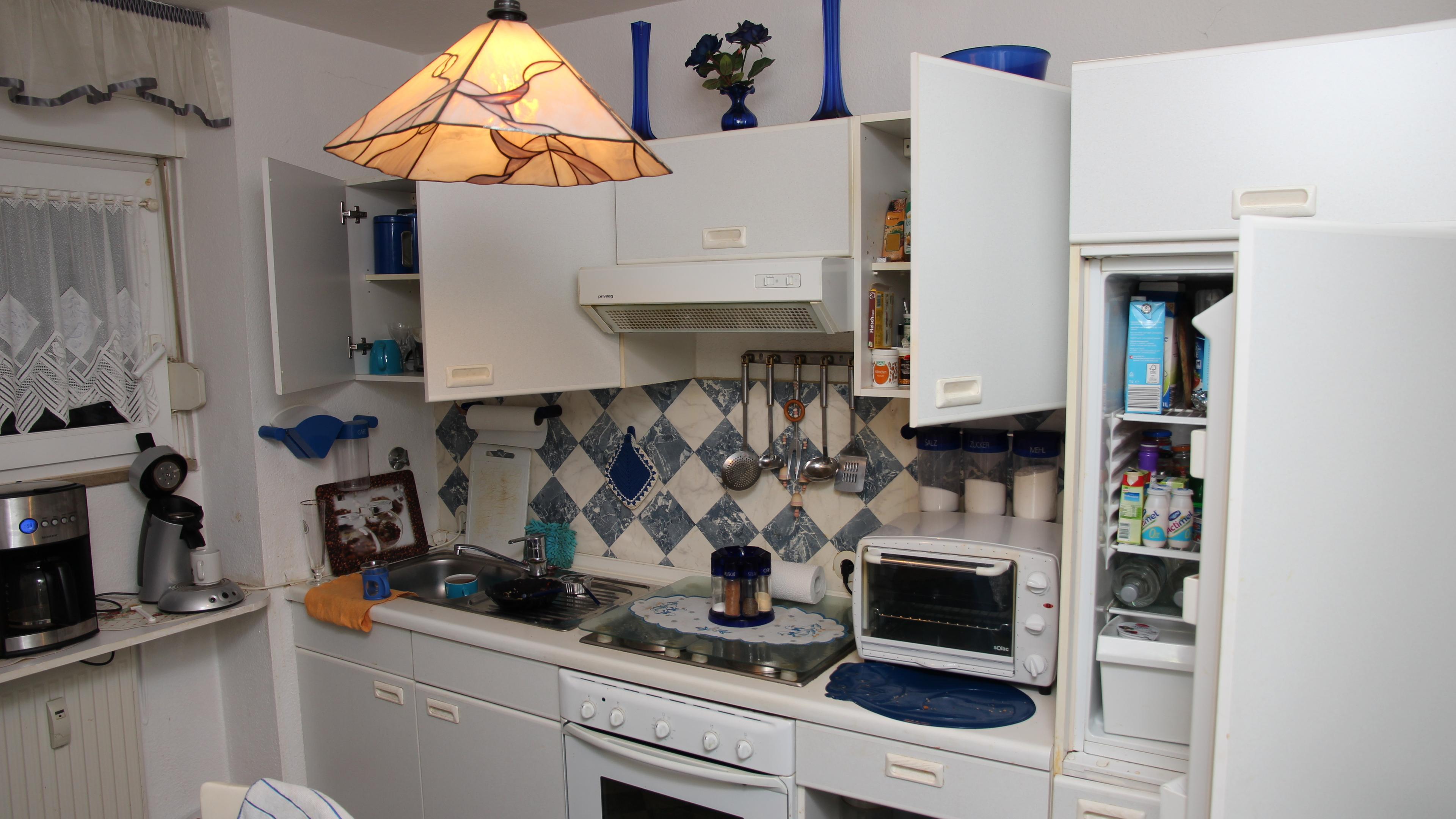  In einer weißen Einbauküche, die sehr aufgeräumt wirkt, sind Ober- und Unterschränke sind teilweise geöffnet. Im geöffneten Kühlschrank an der Seite sieht man verschiedene Getränke und Joghurtpackungen.