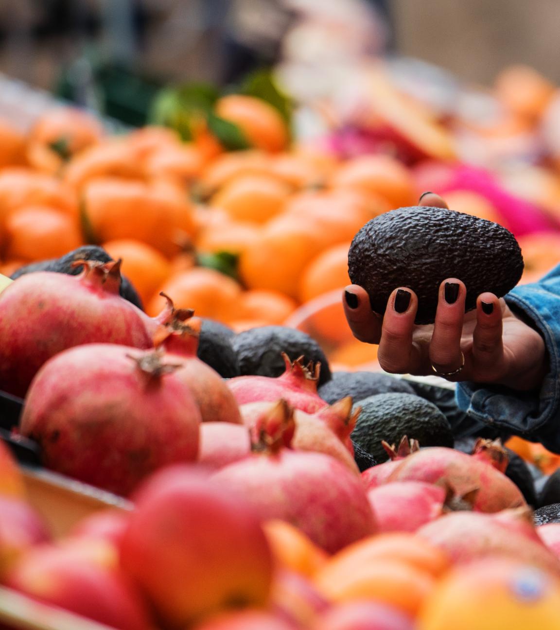 Eine Frau hält eine Avocado in der Hand, während sie an einem Marktstand für Obst und Gemüse steht.