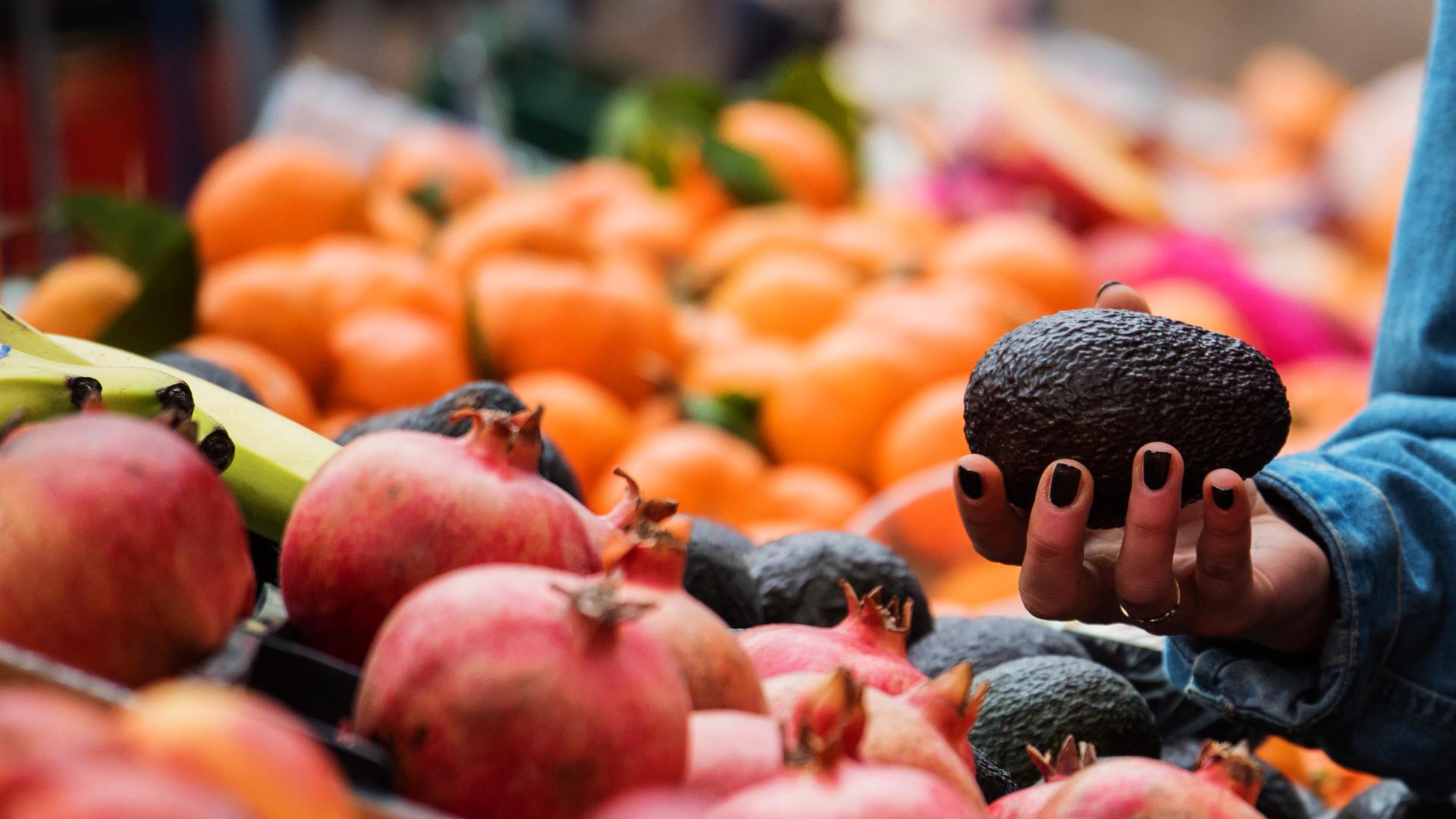 Eine Frau hält eine Avocado in der Hand, während sie an einem Marktstand für Obst und Gemüse steht.