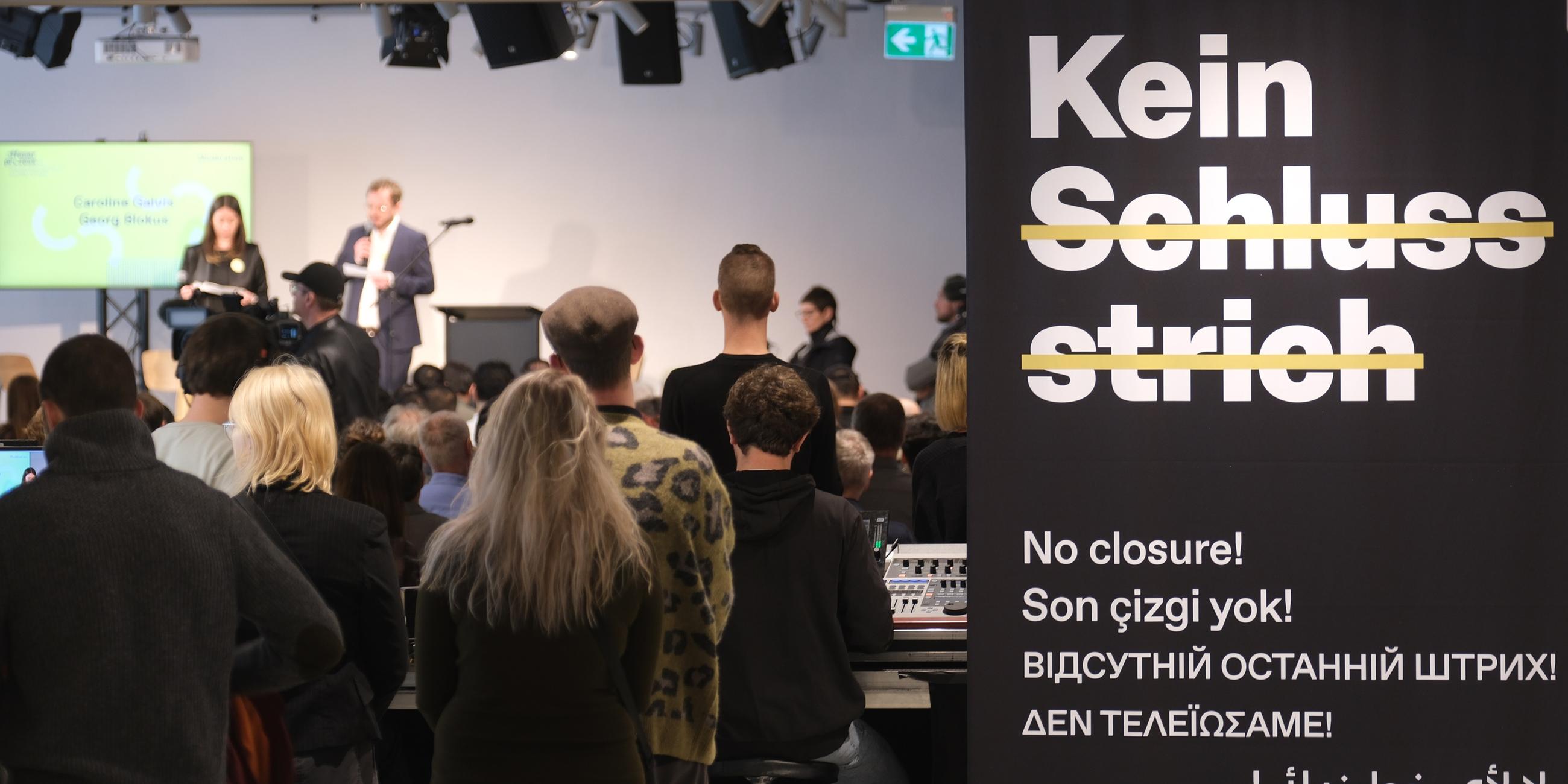 Die Eröffung des Dokumentationszentrums. Zwei Menschen sprechen auf einer Bühne, viele andere Menschen stehen davor. Im rechten Bildrand sind Ausschnitte der Ausstellung zu sehen.