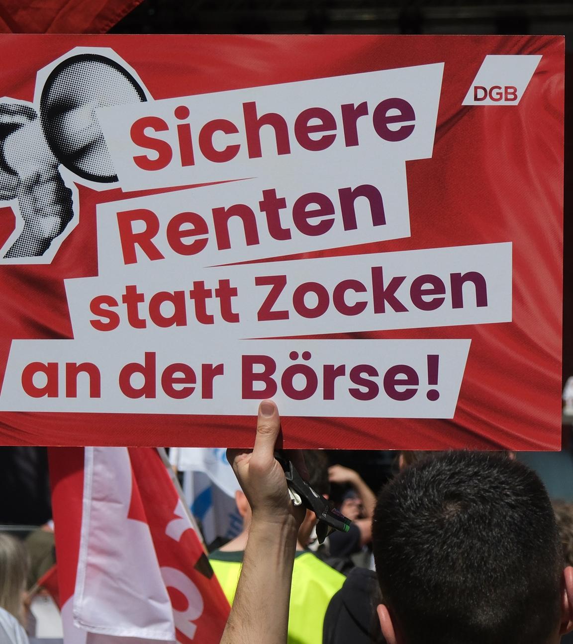 Ein Transparent mit dem Aufdruck "Sichere Renten statt Zocken an der Börse!" und Fahnen werden bei einer Veranstaltung des DGB zum ersten Mai in die Höhe gehalten. 