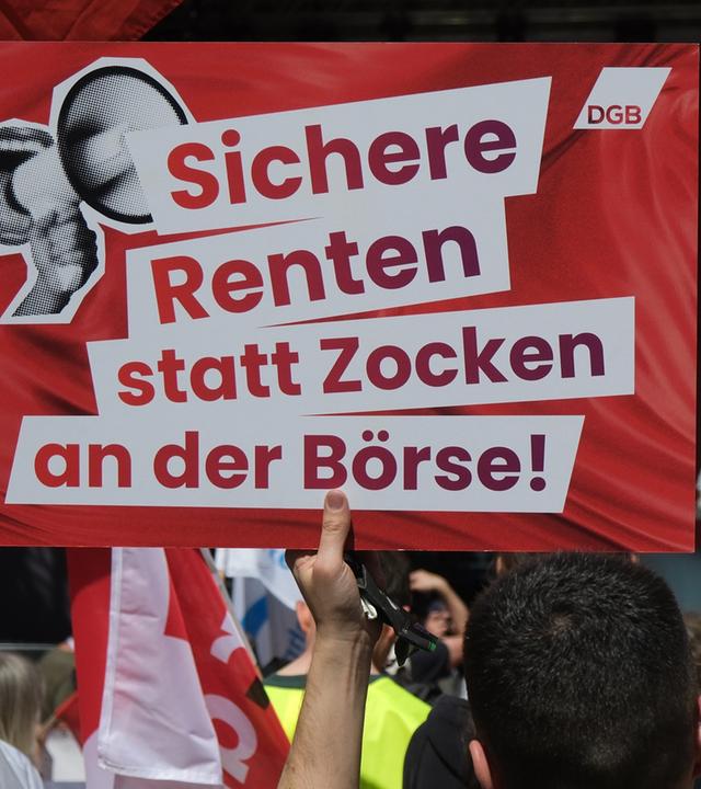Ein Transparent mit dem Aufdruck "Sichere Renten statt Zocken an der Börse!" und Fahnen werden bei einer Veranstaltung des DGB zum ersten Mai in die Höhe gehalten. 