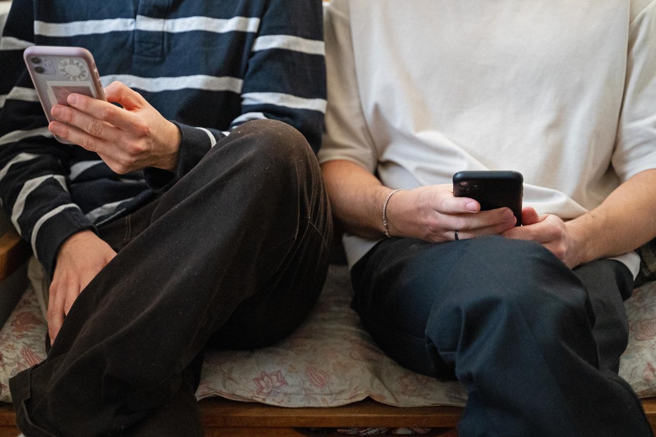 Zwei junge Männer sitzen nebeneinander auf einer Bank und halten ihre Smartphones in den Händen.