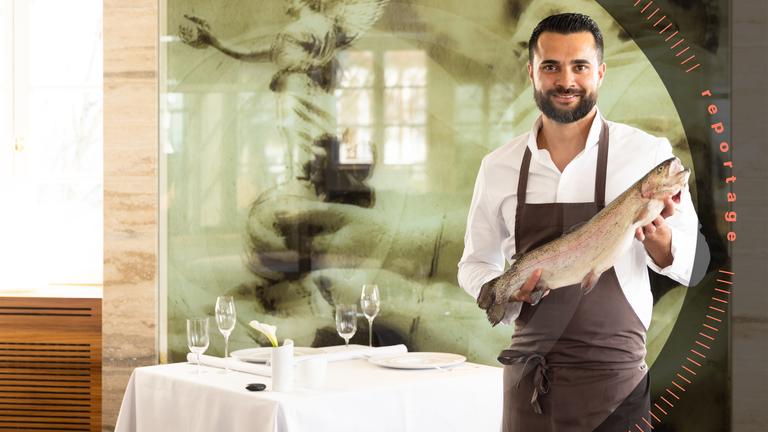 Küchenchef Dennis Kuckuck hält einen großen Fisch in der Hand und lächelt in die Kamera. Er trägt eine Schürze. Im Hintergrund ist der schick gedeckte Tisch eines Restaurants zu sehen.