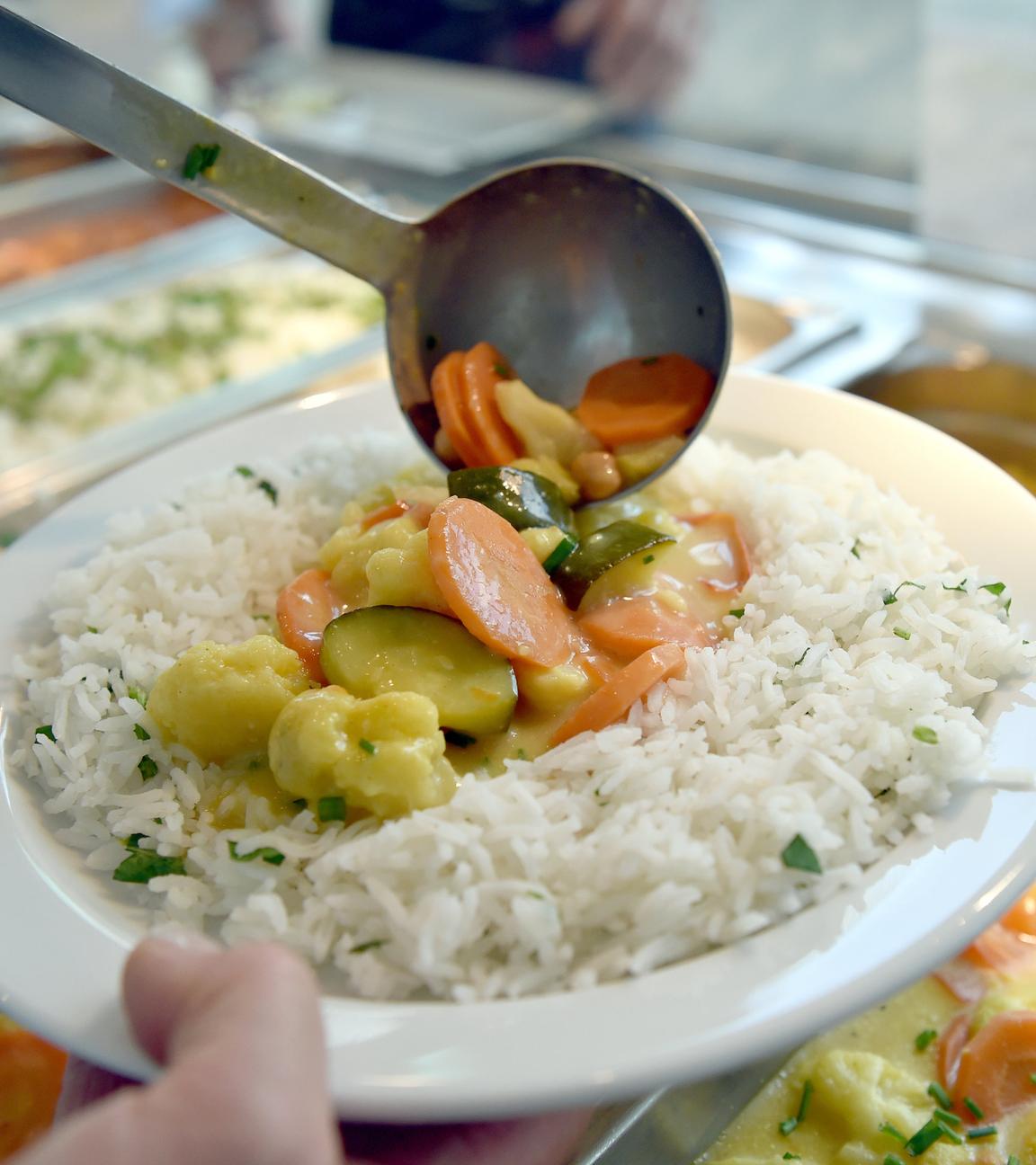 An einem Buffet einer Kantine hält jemand in der einen Hand einen Teller mit Reis und Gemüse und fügt mit der anderen dem Teller eine Kelle Gemüse hinzu.