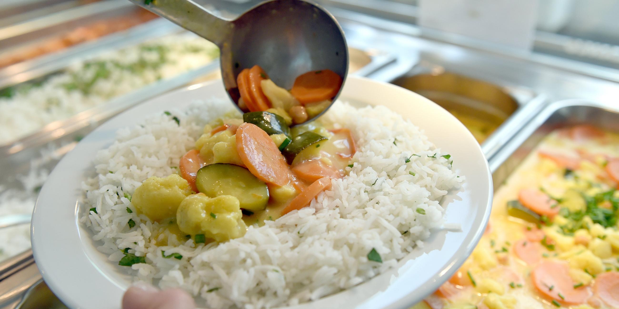 An einem Buffet einer Kantine hält jemand in der einen Hand einen Teller mit Reis und Gemüse und fügt mit der anderen dem Teller eine Kelle Gemüse hinzu.