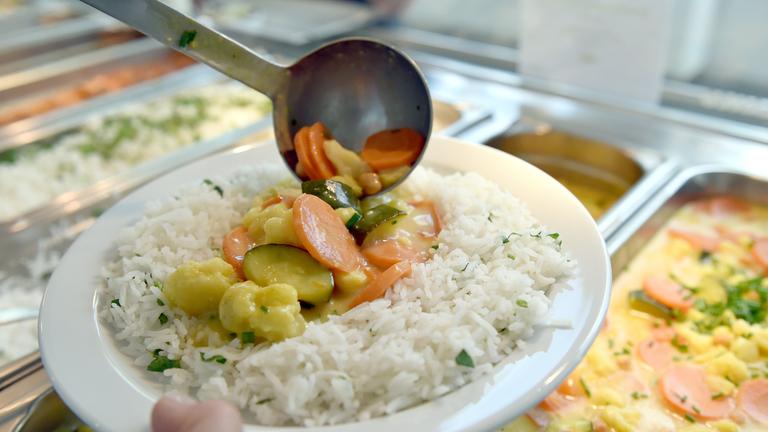 An einem Buffet einer Kantine hält jemand in der einen Hand einen Teller mit Reis und Gemüse und fügt mit der anderen dem Teller eine Kelle Gemüse hinzu.