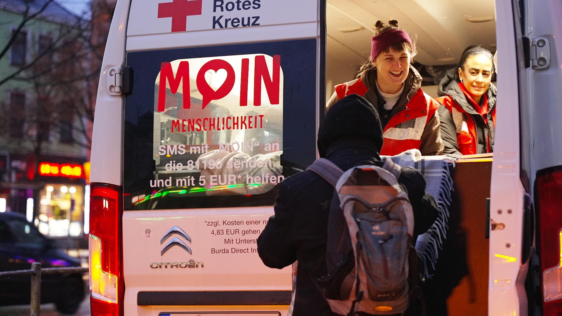 Eine Person reicht aus einem DRK-Auto mit der Aufschrift "Moin Menschlichkeit" etwas an eine Person.