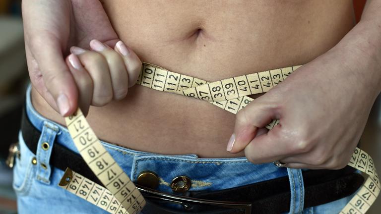 Eine junge Frau legt ein Maßband um ihren Bauch.