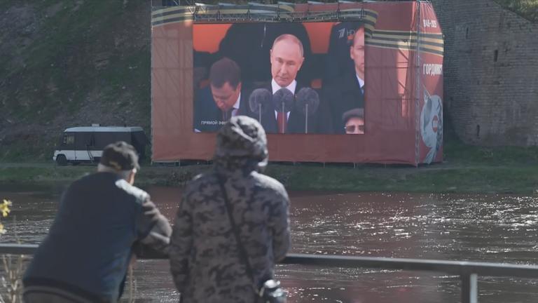 Zwei Personen sehen auf einer großen Leinwand am anderen Ufer eines Flusses eine Rede von Putin