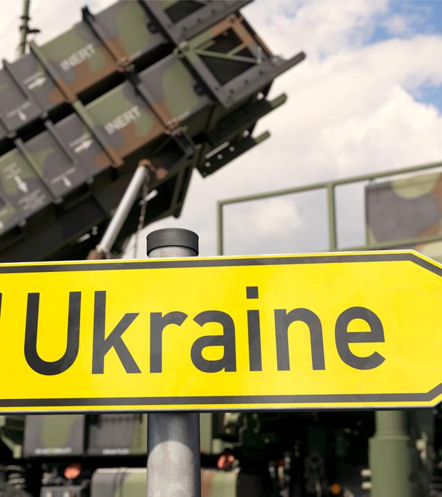 ein Symbolbild, bei dem ein Schild, auf dem "Ukraine" steht, vor militärischen Fahrzeugen zu sehen ist.