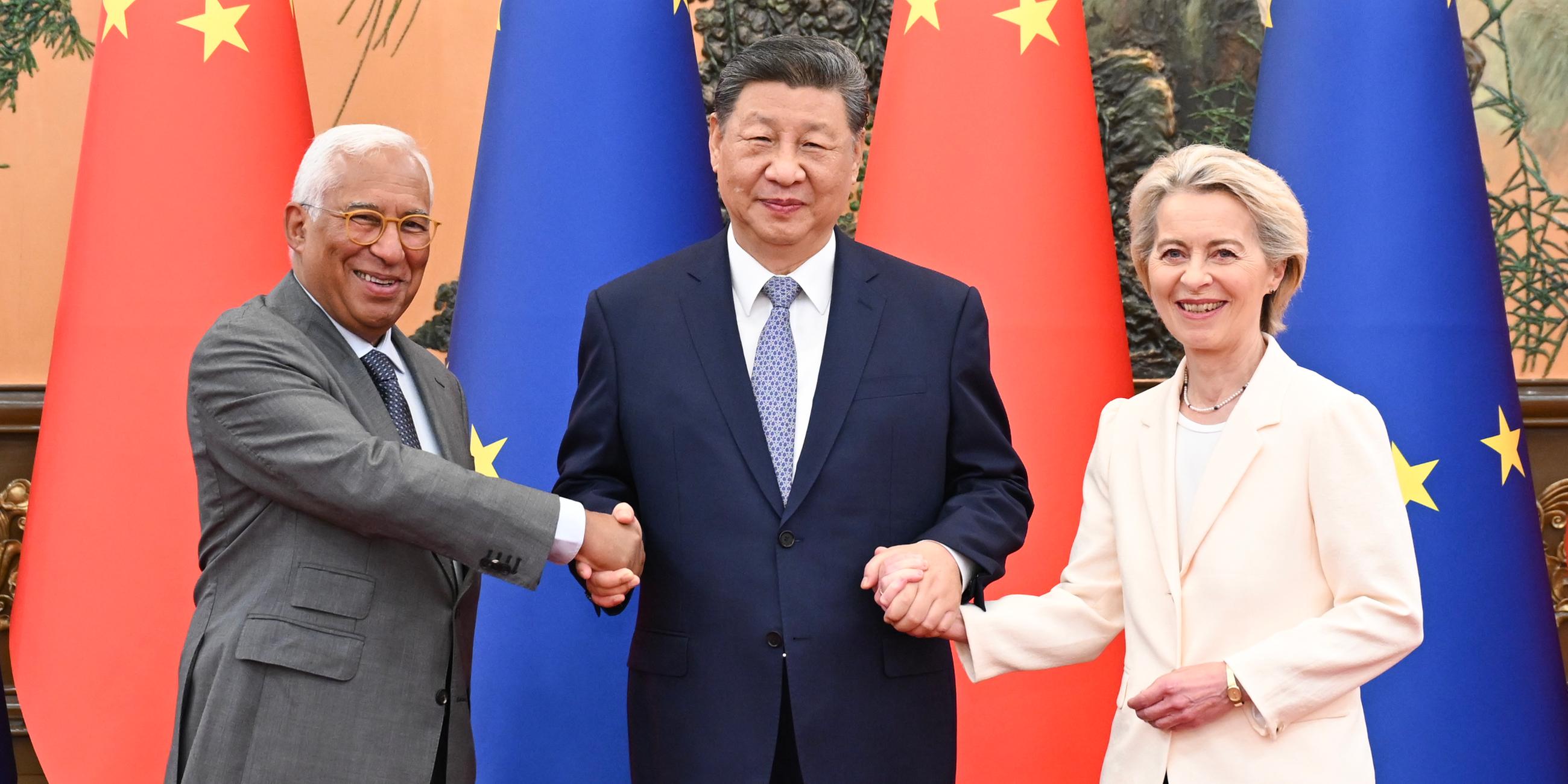 Links Antonio Costa, mittig Xi Jinping, rechts Ursula von der Leyen, hinter ihnen die Fahnen der EU und Chinas. Die drei lächeln und schütteln die Hände.