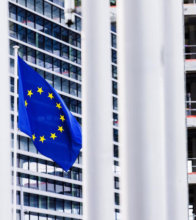 Eine Flagge der Europäischen Union (EU) weht vor dem Gebäude des Europäischen Parlaments und ist zwischen anderen Fahnenmasten hindurch zu sehen. 