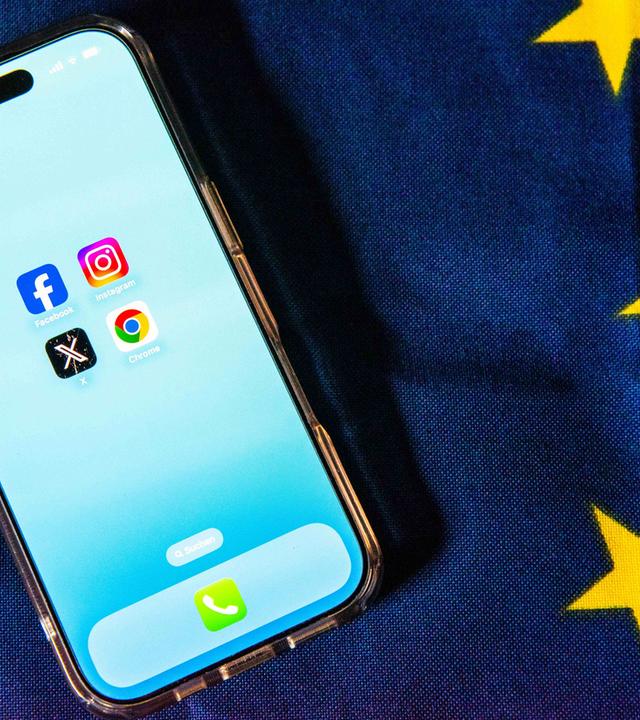 Die Logos von Facebook, Instagram, X und Google Chrome sind auf dem Bildschirm eines Smartphones zu sehen, das auf einer EU-Flagge liegt.
