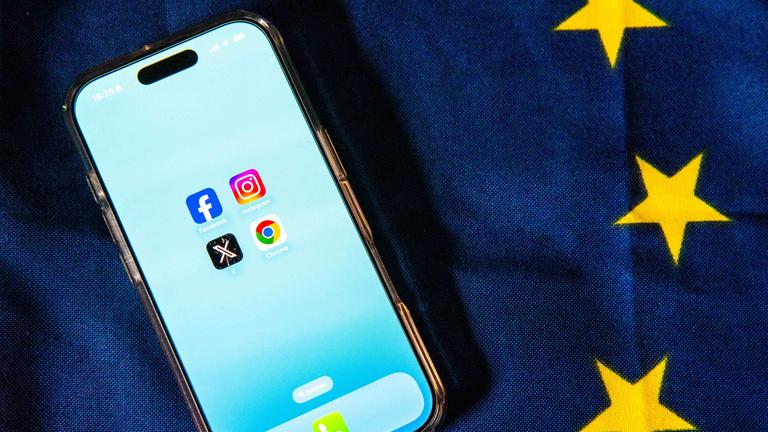Die Logos von Facebook, Instagram, X und Google Chrome sind auf dem Bildschirm eines Smartphones zu sehen, das auf einer EU-Flagge liegt.