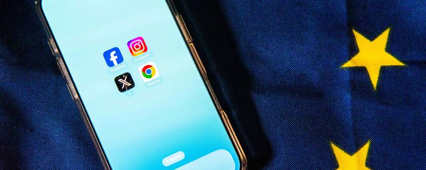 Die Logos von Facebook, Instagram, X und Google Chrome sind auf dem Bildschirm eines Smartphones zu sehen, das auf einer EU-Flagge liegt.