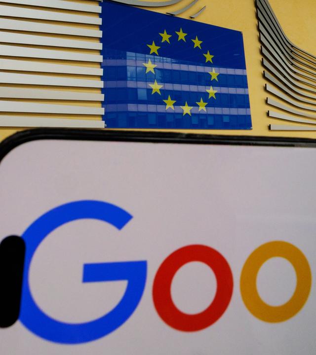 Eine Gesamtansicht zeigt ein Google-Logo auf einem iPhone-Bildschirm vor dem Gebäude der Europäischen Kommission in Brüssel.