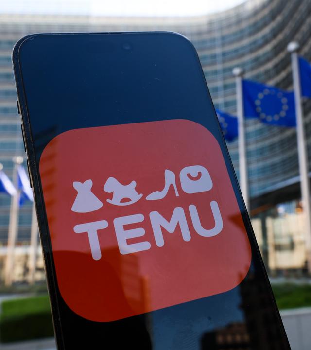 Ein Smartphone mit dem Symbol der "Temu-App" wird vor dem Gebäude der EU-Kommission mit zahlreichen EU-Flaggen davor hochgehalten