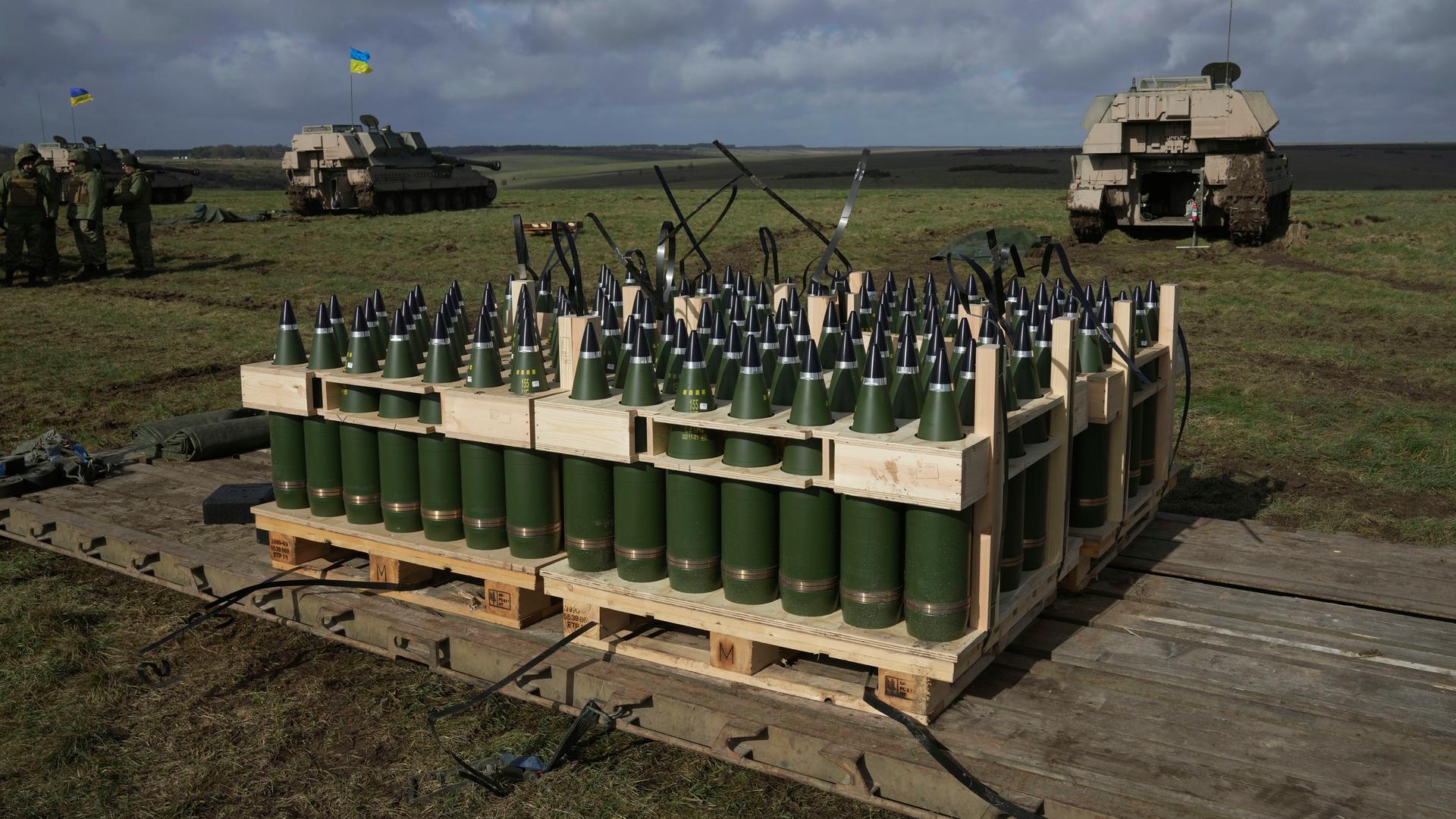 Im Vordergrund steht Artillerie-Munition. Im Hintergrund Militärfahrzeuge mit ukrainischen Flaggen.
