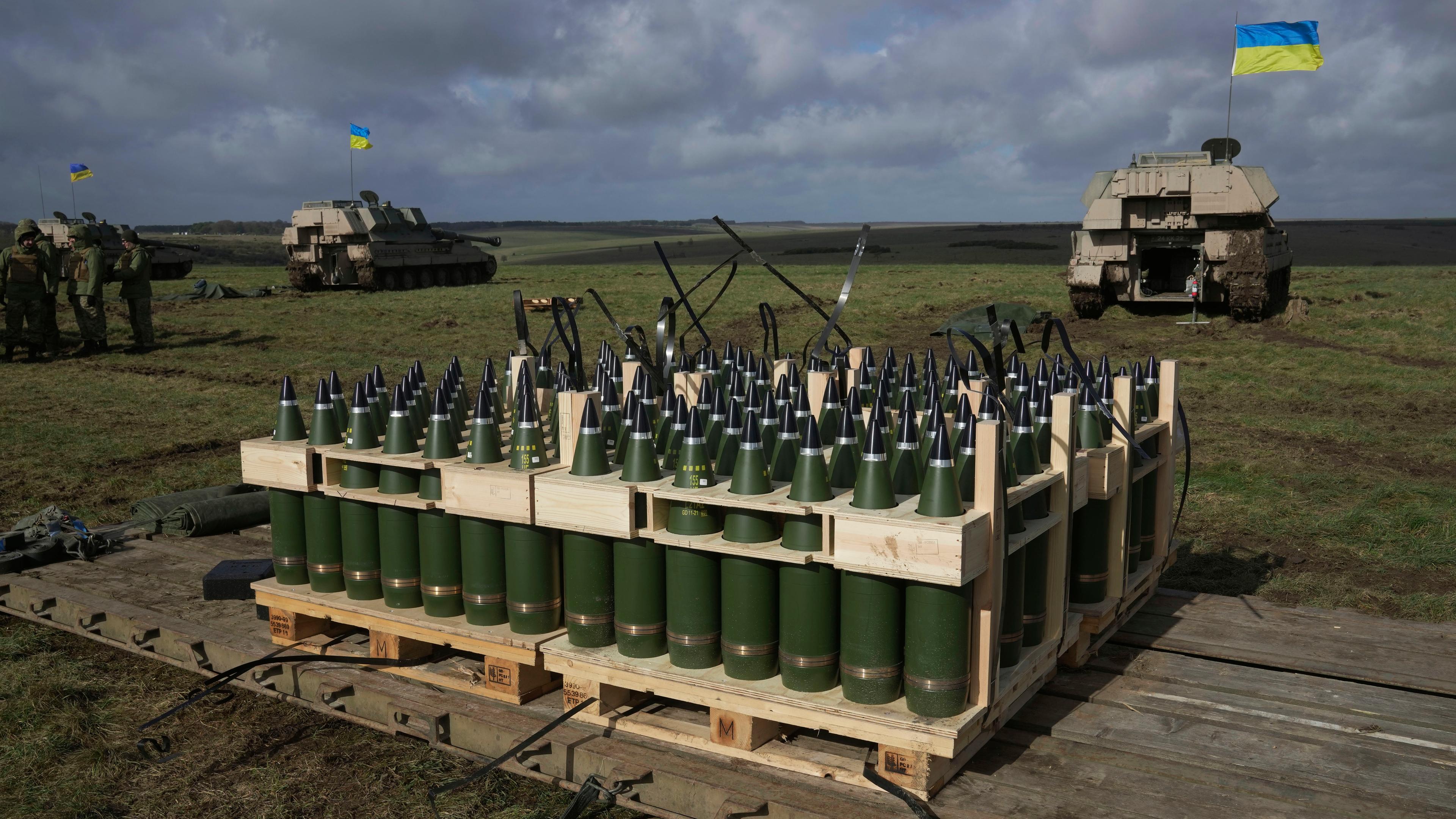 Im Vordergrund steht Artillerie-Munition. Im Hintergrund Militärfahrzeuge mit ukrainischen Flaggen.