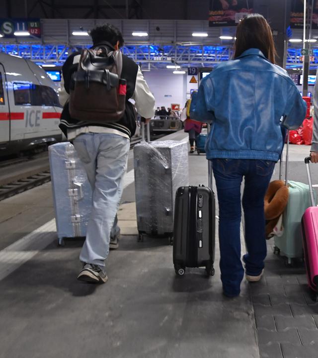 Drei Reisende mit Rucksaecken und zahlreichen Rollkoffern gehen am Bahnsteig entlang eines ICE-Fernzuges im Muenchner Hauptbahnhof.