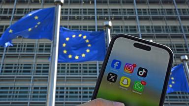 Eine Person hält ein Smartphone mit den Logos der Social-Media-Apps Facebook, Instagram, TikTok, X, Snapchat und WhatsApp vor Fahnen der Europäischen Union und dem Gebäude der Europäischen Kommission.