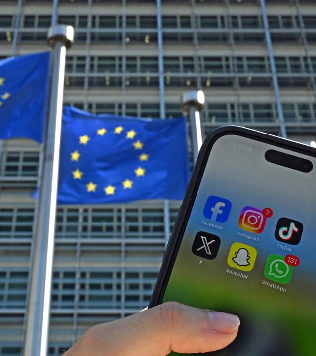 Eine Person hält ein Smartphone mit den Logos der Social-Media-Apps Facebook, Instagram, TikTok, X, Snapchat und WhatsApp vor Fahnen der Europäischen Union und dem Gebäude der Europäischen Kommission.