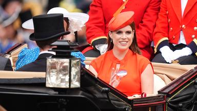 Auf dem Bild ist Prinzessin Eugenie zu sehen. Diese sitzt in einer offenen schwarze Kutsche. Sie trägt ein leuchtenden orangefarbenes Kleid mit passendem Hut.