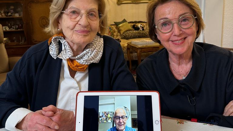 Eva Szepesi (links) und Eva Umlauf sitzen lächelnd nebeneinander an einem Tisch. Vor ihnen steht ein Tablet, auf dem Naftali Fürst zu sehen ist.