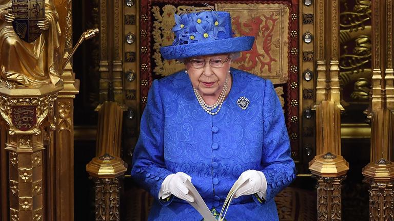Das Bild zeigt Queen Elizabeth II mit einem blauen Hut mit gelben Blüten