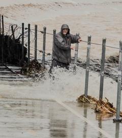 EIn Mann stapft entlang einer teils überfluteten Straße und hält sich dabei an einem Geländer fest.