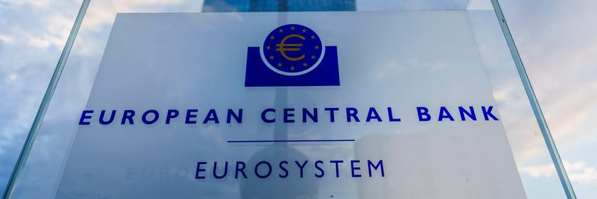 Ein Schild mit der Aufschrift "European Central Bank - Eurosystem". Im Hintergrund die Zentrale der EZB.