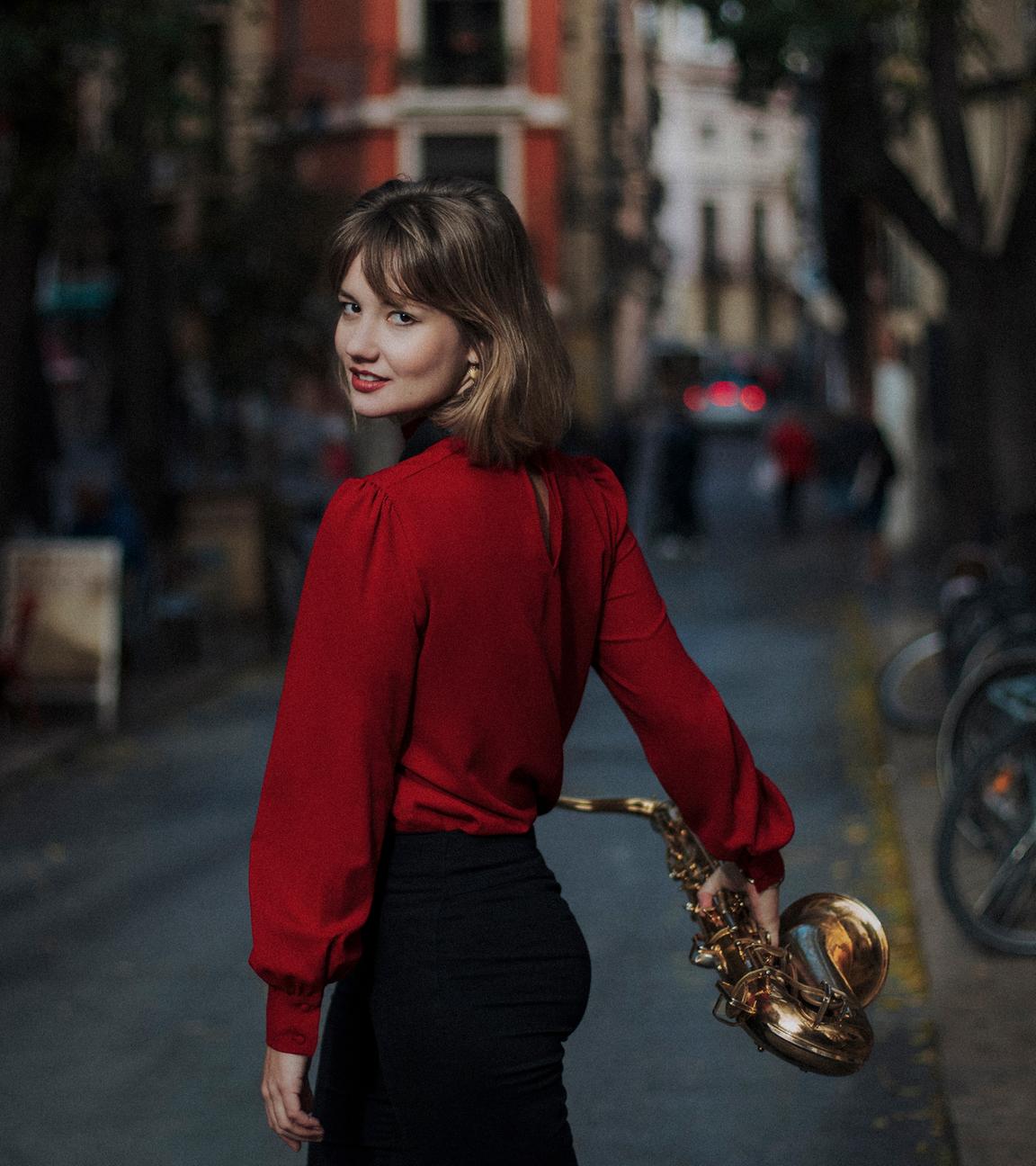 Die junge Saxophonistin Fabia Mantwill in roter Bluse und schwarzer Hose, von der Kamera auf der Straße in einer Wohngegend weglaufend, sich umdrehend in der Bewegung und das Saxophon in der rechten Hand habend.